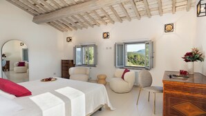 Un dormitorio con cama, dos sillas, cómoda, espejo y dos ventanas con vistas a zonas verdes.