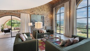 Un salón amplio con una pared de piedra curva, ventanas grandes que ofrecen vistas al campo, un juego de sofá verde y una mesa de centro de vidrio.