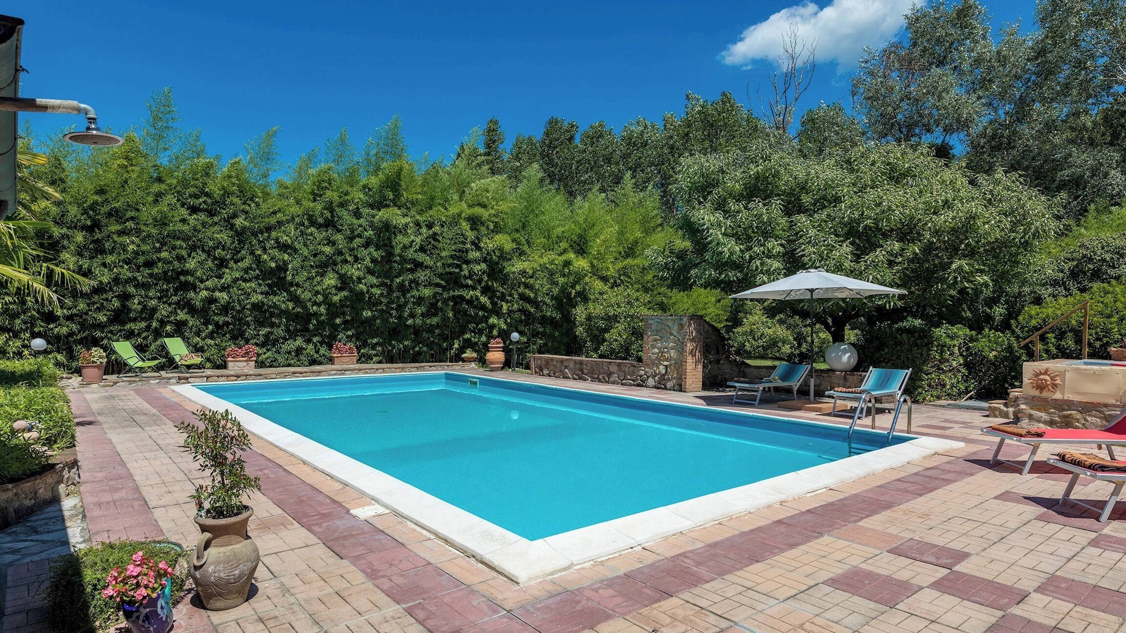 A rectangular swimming pool with a tiled edge, surrounded by a brick patio. There are lounge chairs and a small umbrella providing shade.