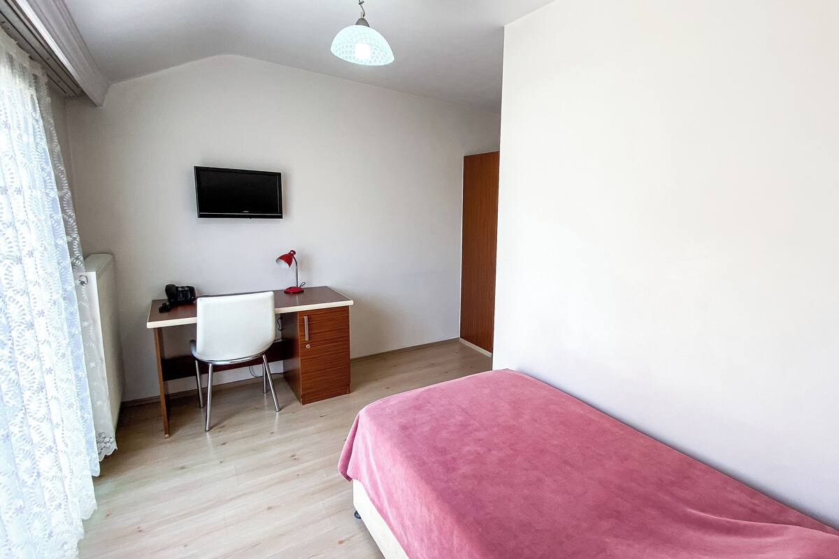 A bedroom with a single bed, a desk with a chair, a television on the wall, and a wooden door.
