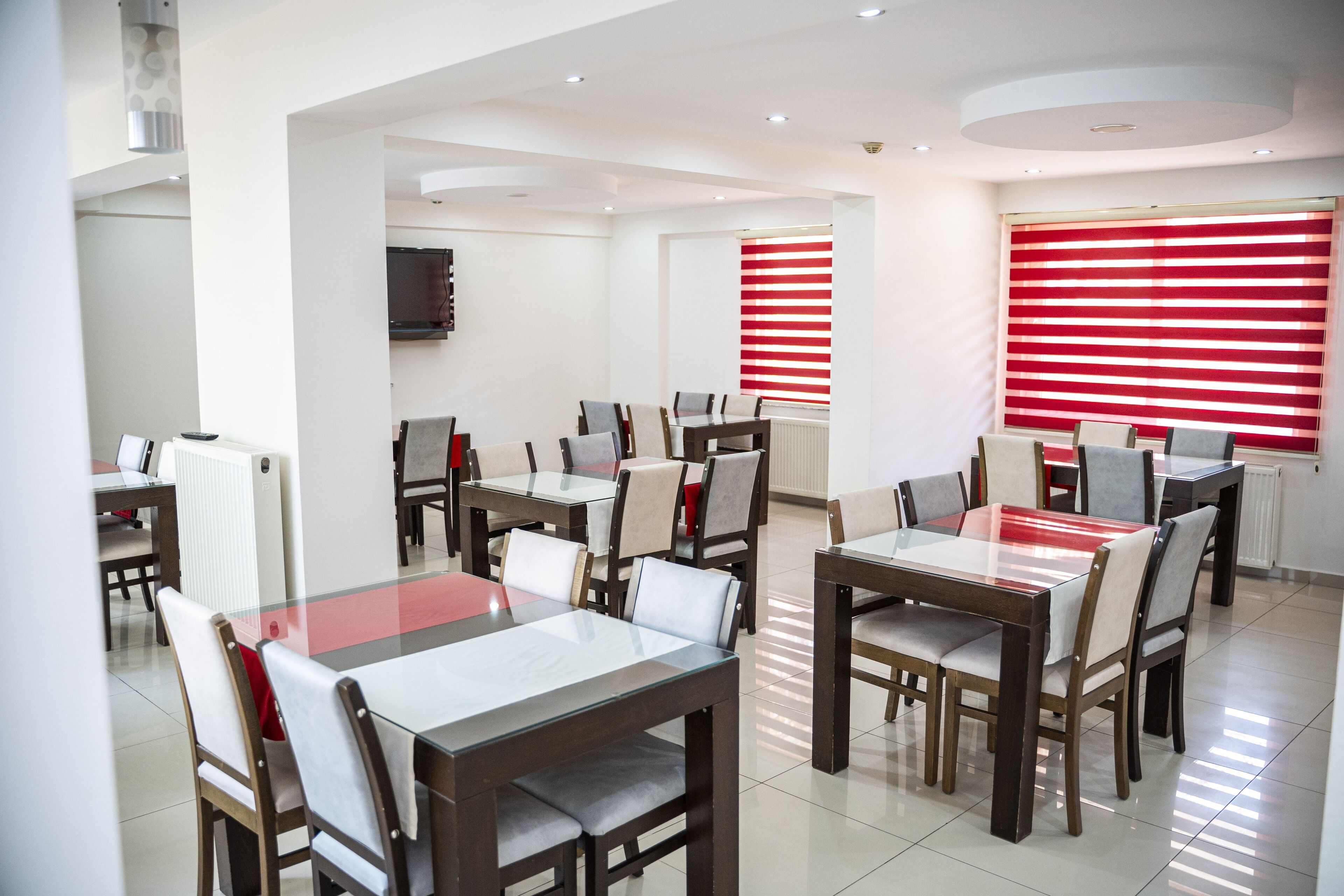 A modern dining area with multiple tables and chairs, a television mounted on the wall, and red-striped window blinds.