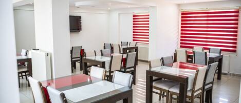 A modern dining area with multiple tables and chairs, a television mounted on the wall, and red-striped window blinds.