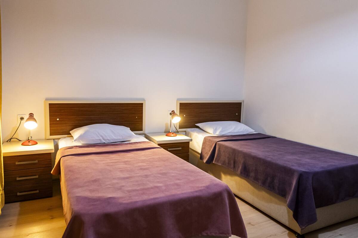A hotel room with two single beds, each with a wooden headboard, a bedside table, and a lamp.