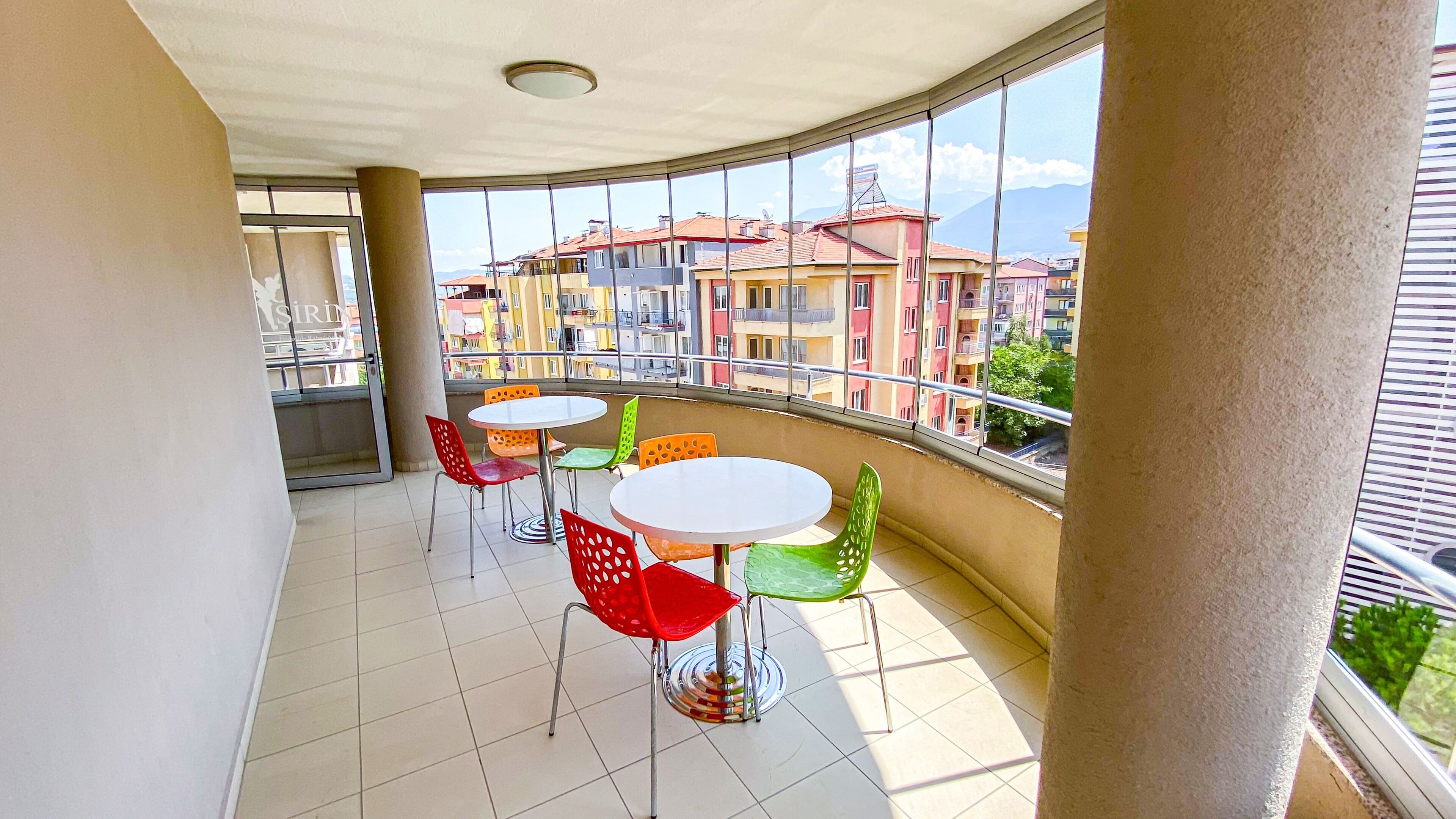 Varanda com mesa branca e quatro cadeiras coloridas, com vista para área residencial com edifícios coloridos e uma montanha ao fundo.