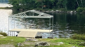Una estructura metálica con techo, posiblemente un pabellón, cerca de un cuerpo de agua con un bosque al fondo.