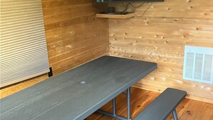 Una cabaña de madera con una mesa de picnic metálica y un banco, un televisor montado y una ventana con persianas.