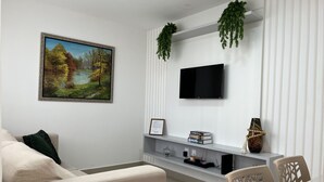 Ett modernt vardagsrum med en beige soffa, en platt-TV som är monterad på en vit vägg, en inramad tavla och en liten hylla med böcker och dekorativa föremål.