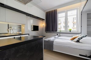 Una habitación de hotel moderna con zona de cocina, una cama con almohadas, una ventana con cortinas y un televisor de pantalla plana montado en la pared.