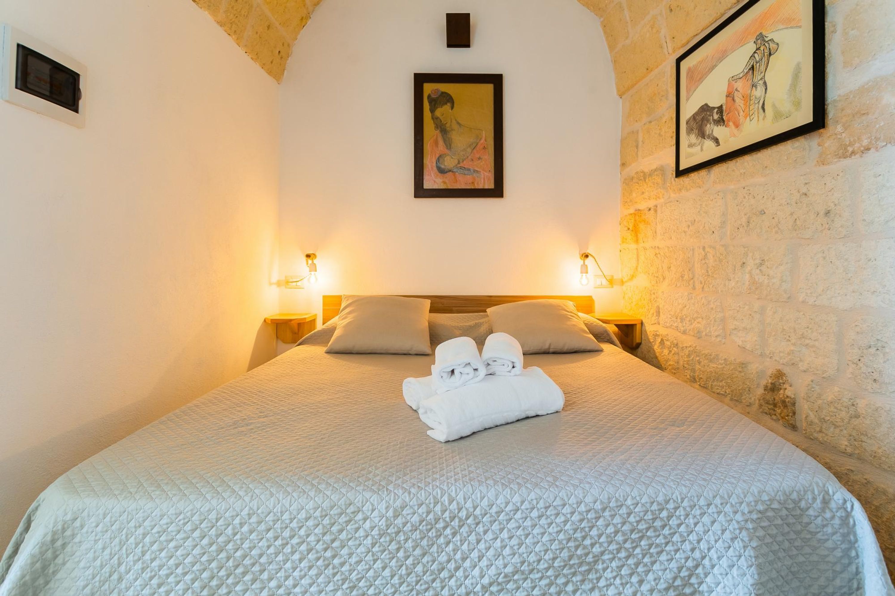 A neatly made bed with a quilted white bedspread and two pillows, a framed painting above, and two wall-mounted lamps on either side.