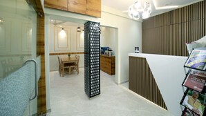 Un interior moderno con techo de madera, una columna central con diseño decorativo, una zona de comedor con mesa y sillas de madera, y un mostrador de recepción con revistas.