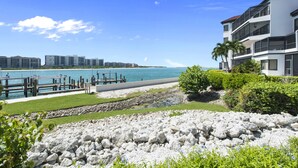 A beachfront property with a rocky shoreline, a wooden dock, and a white building with balconies overlooking the ocean. - Special Savings January 2024 31 days minimum! (Naples)