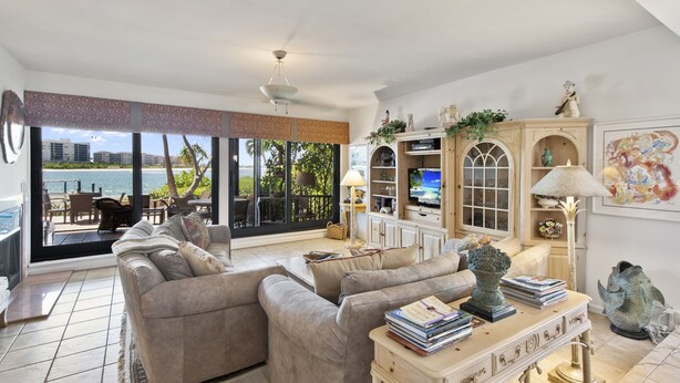 A spacious living room with a large window overlooking a balcony, a sofa set, a television, and a decorative cabinet. - Special Savings January 2024 31 days minimum! (Naples)