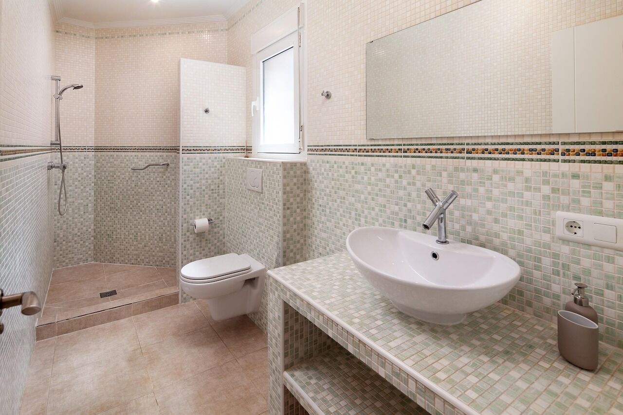 Un baño con ducha, inodoro y lavamanos, que presenta paredes de mosaicos y una encimera blanca.