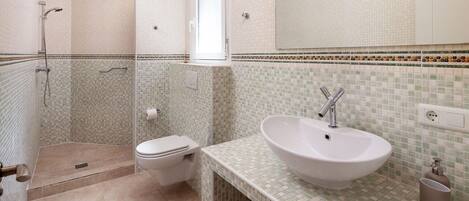 Un baño con ducha, inodoro y lavamanos, que presenta paredes de mosaicos y una encimera blanca.