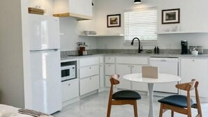 A modern kitchen with white cabinets, a white refrigerator, a dining table with two chairs, and a window with blinds. - Chic studio near ACU (Abilene)
