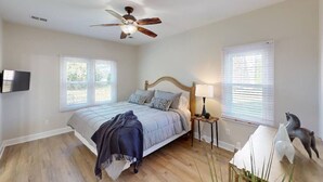 A bedroom with a bed, a ceiling fan, a TV, two windows, a nightstand, and a decorative horse statue.