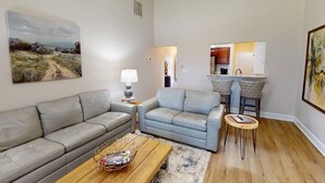 A living room with a grey sofa, a blue armchair, a wooden coffee table, and a small side table with a lamp. There is a painting on the wall and a kitchen area in the background.