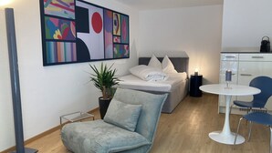 Ein modernes Hotelzimmer mit Bett, einer Sitzecke mit Stuhl und kleinem Tisch, einer Topfpflanze und einem großen abstrakten Gemälde an der Wand.