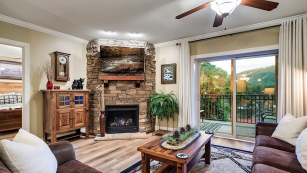 A living room with a stone fireplace, a ceiling fan, and a view of a balcony with a railing. - Downtown Gatlinburg Condo | Walk to Attractions! (Gatlinburg)