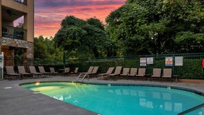 A swimming pool with lounge chairs and a stone wall, surrounded by trees and a sunset sky. - Downtown Gatlinburg Condo | Walk to Attractions! (Gatlinburg)