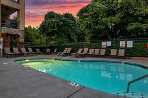 A swimming pool with lounge chairs and a stone wall, surrounded by trees and a sunset sky. - Downtown Gatlinburg Condo | Walk to Attractions! (Gatlinburg)