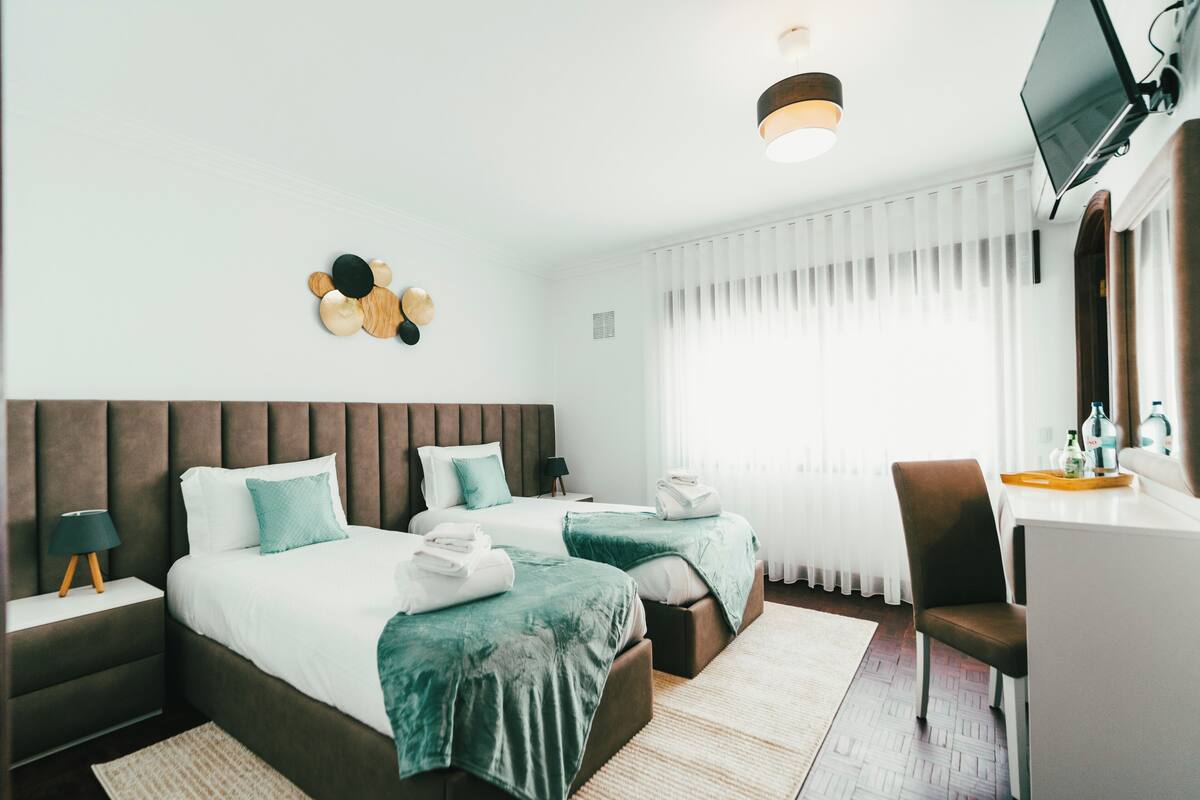 A hotel room with two beds, a desk, a chair, a TV, and a large window with curtains.