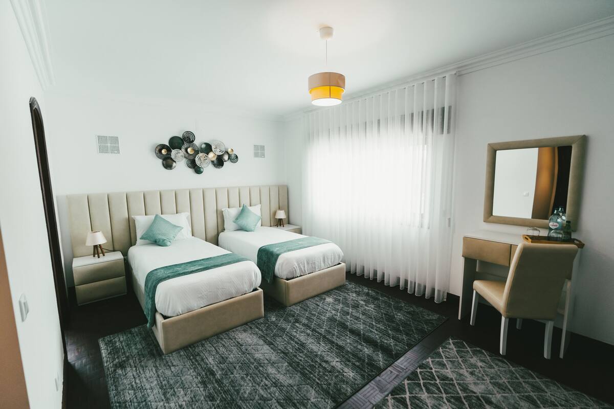 A hotel room with two beds, a desk, a chair, a mirror, and a window with curtains.