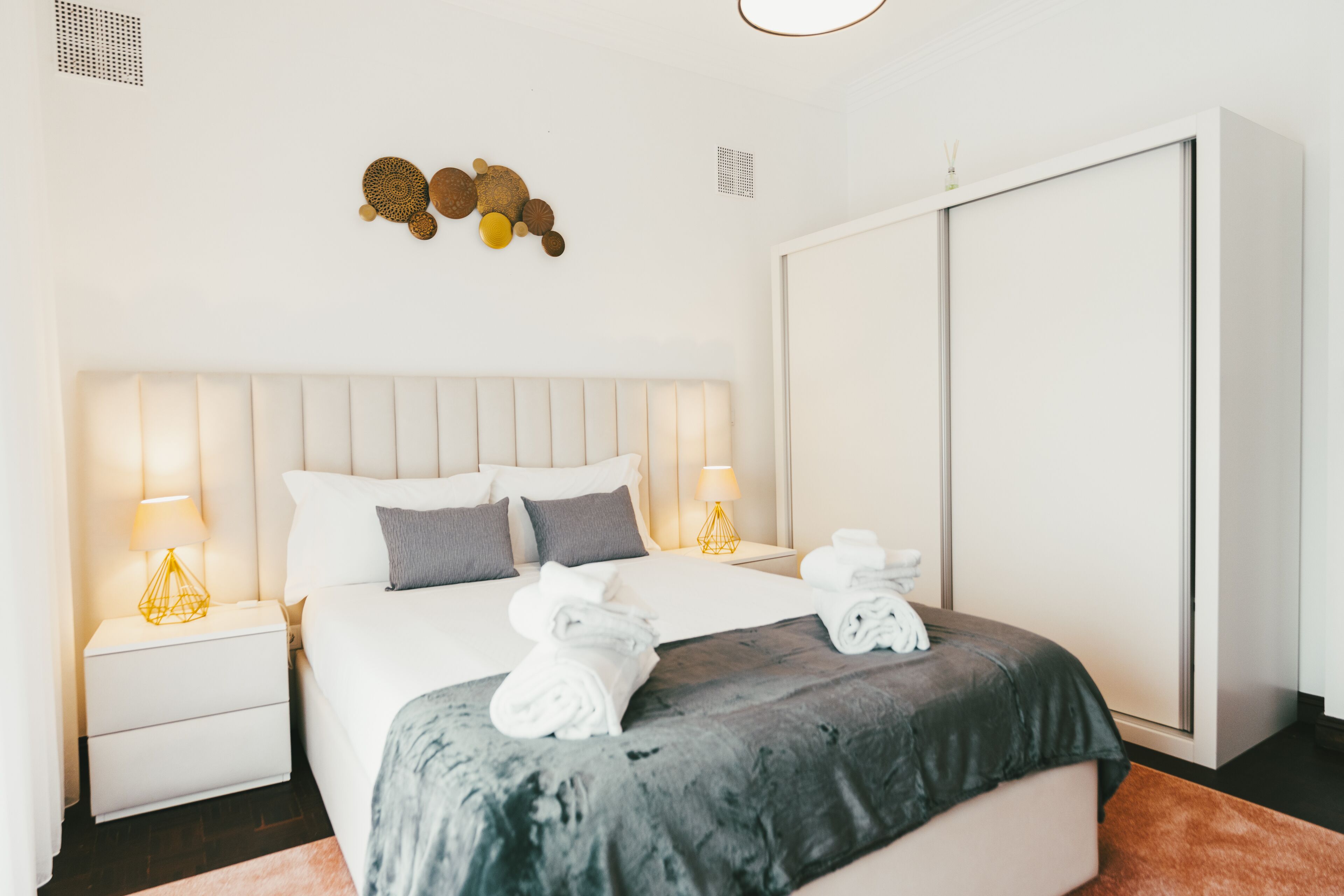A modern hotel room with a large bed, two bedside tables with lamps, a wardrobe, and decorative wall art.