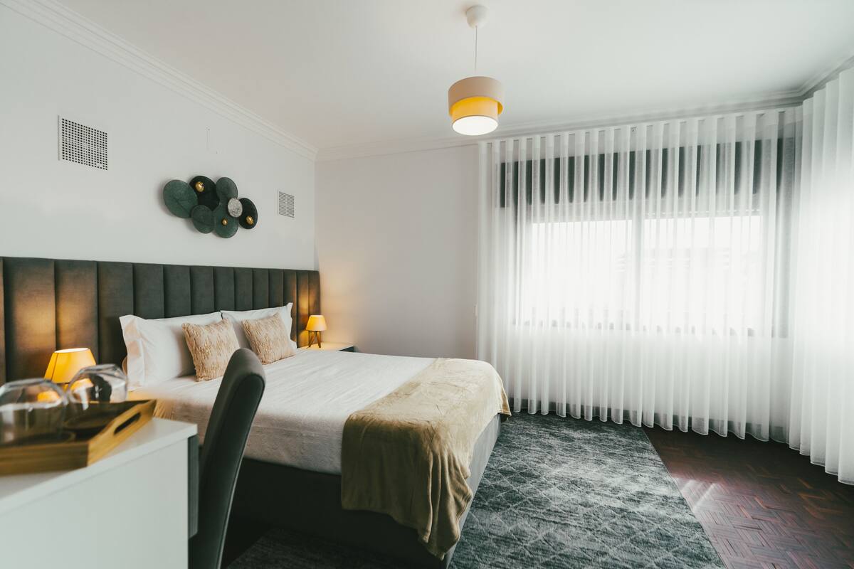 A neatly arranged hotel room with a bed, bedside table, lamp, and a large window with sheer curtains.