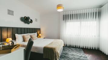 Et pænt indrettet hotelværelse med seng, natbord, lampe og et stort vindue med let gardin.