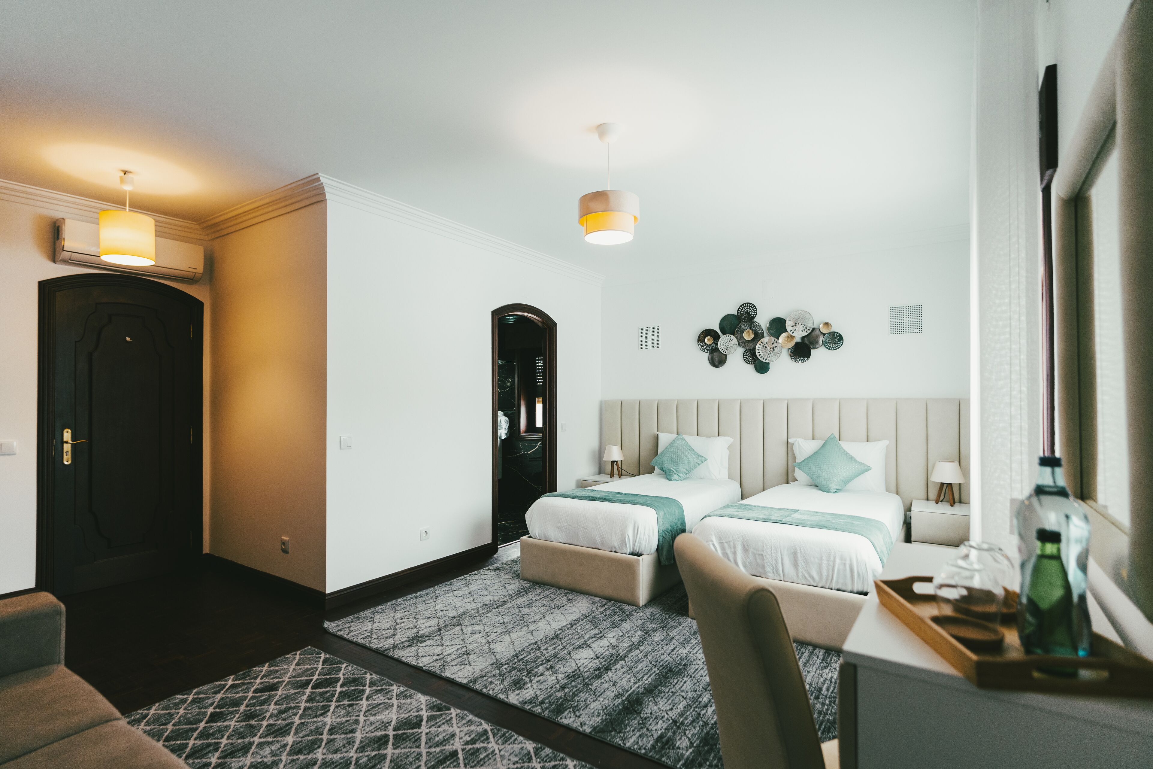 Ett hotellrum med två sängar, ett skrivbord med en flaska och glas, en stol, en mönstrad matta, en väggklocka och en taklampa.