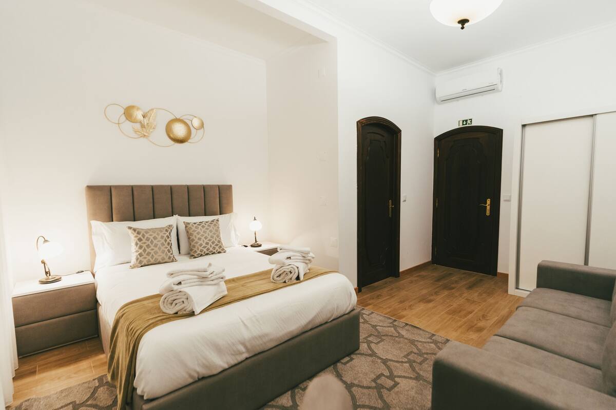 A hotel room with a bed, bedside tables, a desk, a chair, a wardrobe, and an air conditioning unit.