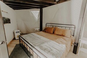 Un dormitorio con estructura de cama metálica, cubrecama estampado y dos almohadas. Hay una mesita de noche pequeña y un calentador blanco en el suelo.
