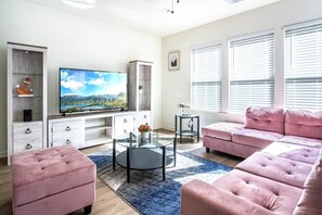 A modern living room with a flat-screen TV, a pink sofa set, a glass coffee table, and a decorative plant.