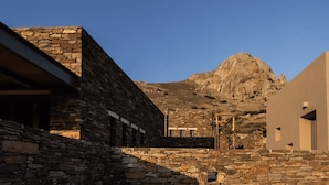 A stone building with a modern structure in the background, a mountain, and a clear sky. - Under the Linden Tree Tinos (Tinos)
