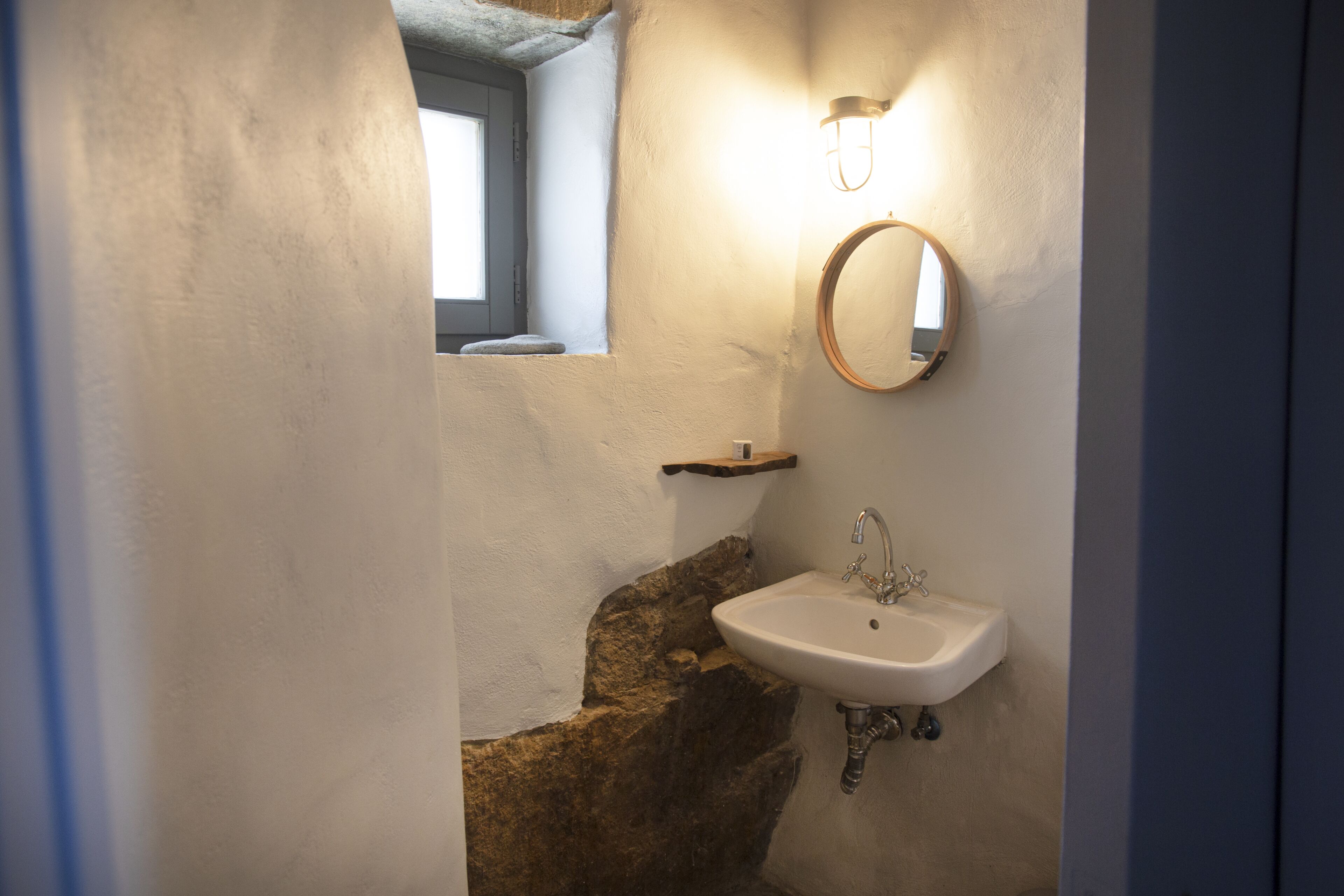 A bathroom with a stone accent wall, a white sink, a round mirror, and a small shelf.