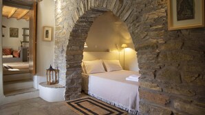 A bedroom with a stone archway, a bed with white linens, a nightstand, a lantern, and a framed picture on the wall. - Under the Linden Tree Tinos (Tinos)
