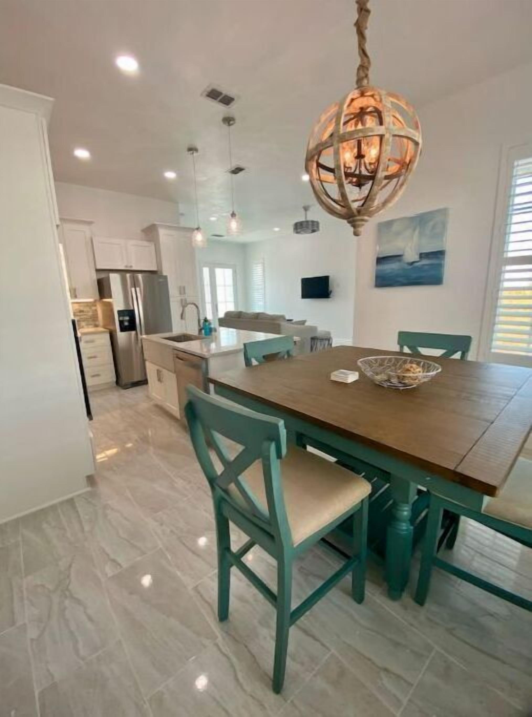 A modern kitchen with white cabinets, a stainless steel refrigerator, and a dining area with a wooden table and teal chairs.