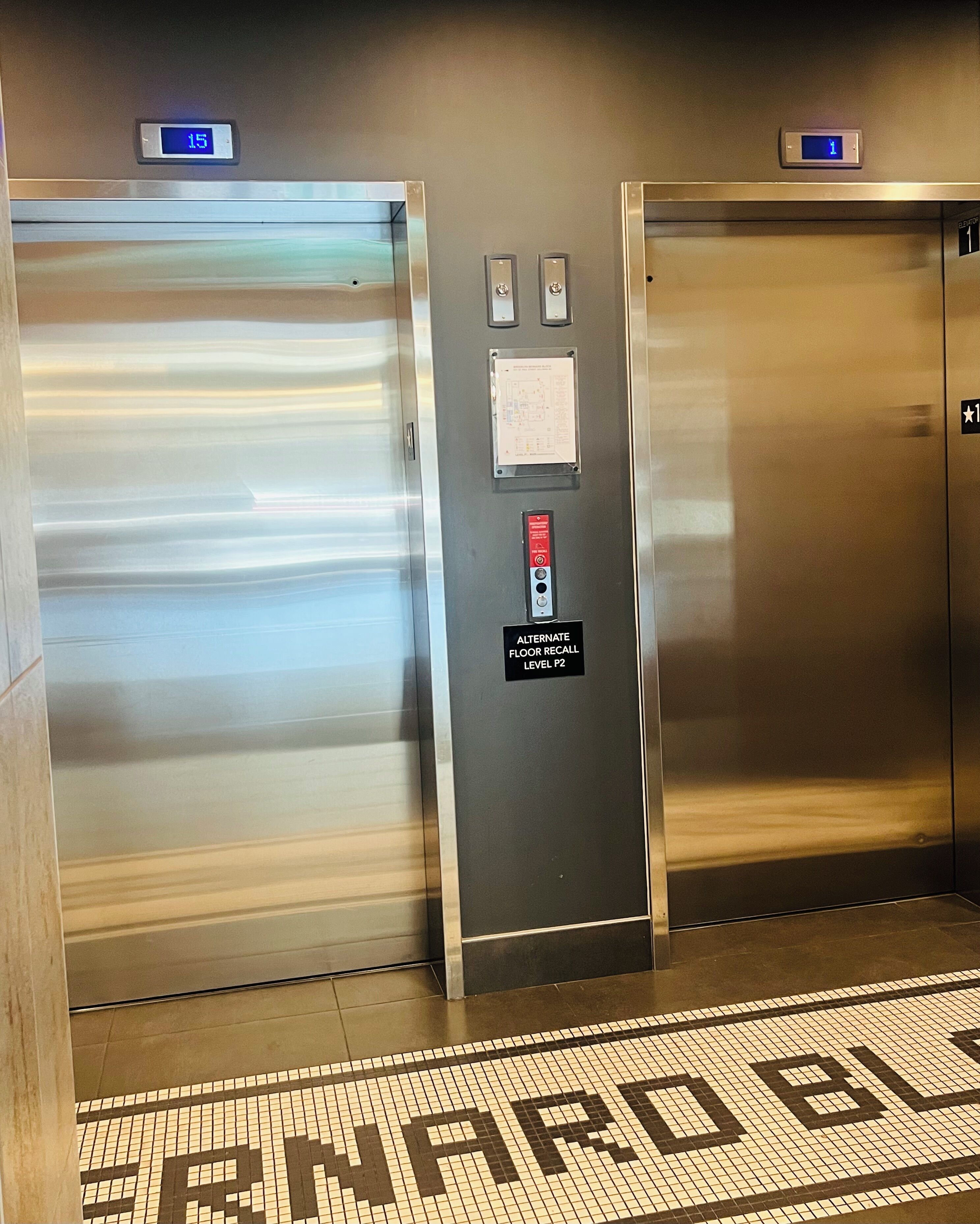 Two elevator doors with digital displays, a floor recall panel, and a tiled floor with a directional arrow.