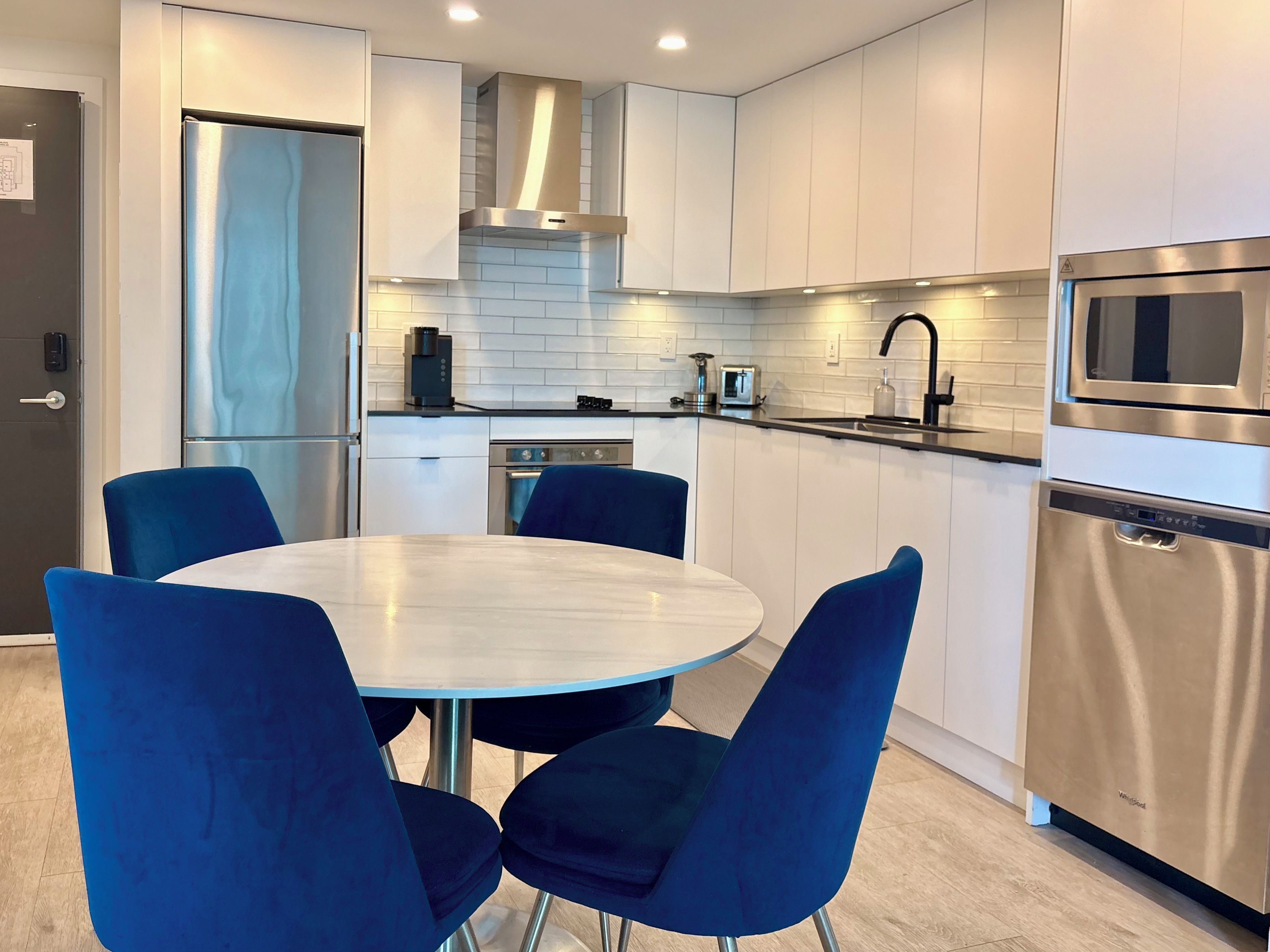 Una cocina moderna con gabinetes blancos, electrodomésticos de acero inoxidable y una mesa redonda con sillas azules.