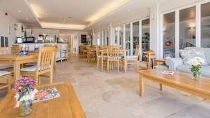 A spacious dining area with wooden tables and chairs, a kitchen in the background, and large windows offering views of the outdoors. - 14 Burgh Island Causeway (Kingsbridge)