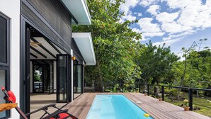 A modern outdoor pool area with a wooden deck, glass doors, and a red lounge chair. - Casa Kali - 4-Min Walk to Beach - AC & Pool (Puerto Viejo de Talamanca)
