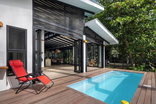 A modern outdoor pool area with a red lounge chair, a covered patio, and a view of the interior.