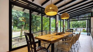 An outdoor dining area with a wooden table and wicker chairs, surrounded by large windows and a view of greenery. - Casa Kali - 4-Min Walk to Beach - AC & Pool (Puerto Viejo de Talamanca)