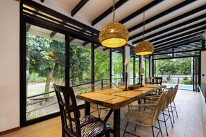 An outdoor dining area with a wooden table and wicker chairs, surrounded by large windows and a view of greenery.