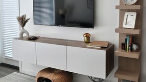 Ein modernes Wohnzimmer mit einem flachbildfernseher an einer weißen Wand, einem hölzernen Konsolentisch mit Dekorationen, einem gewebten Pouf und einem gemusterten Teppich.