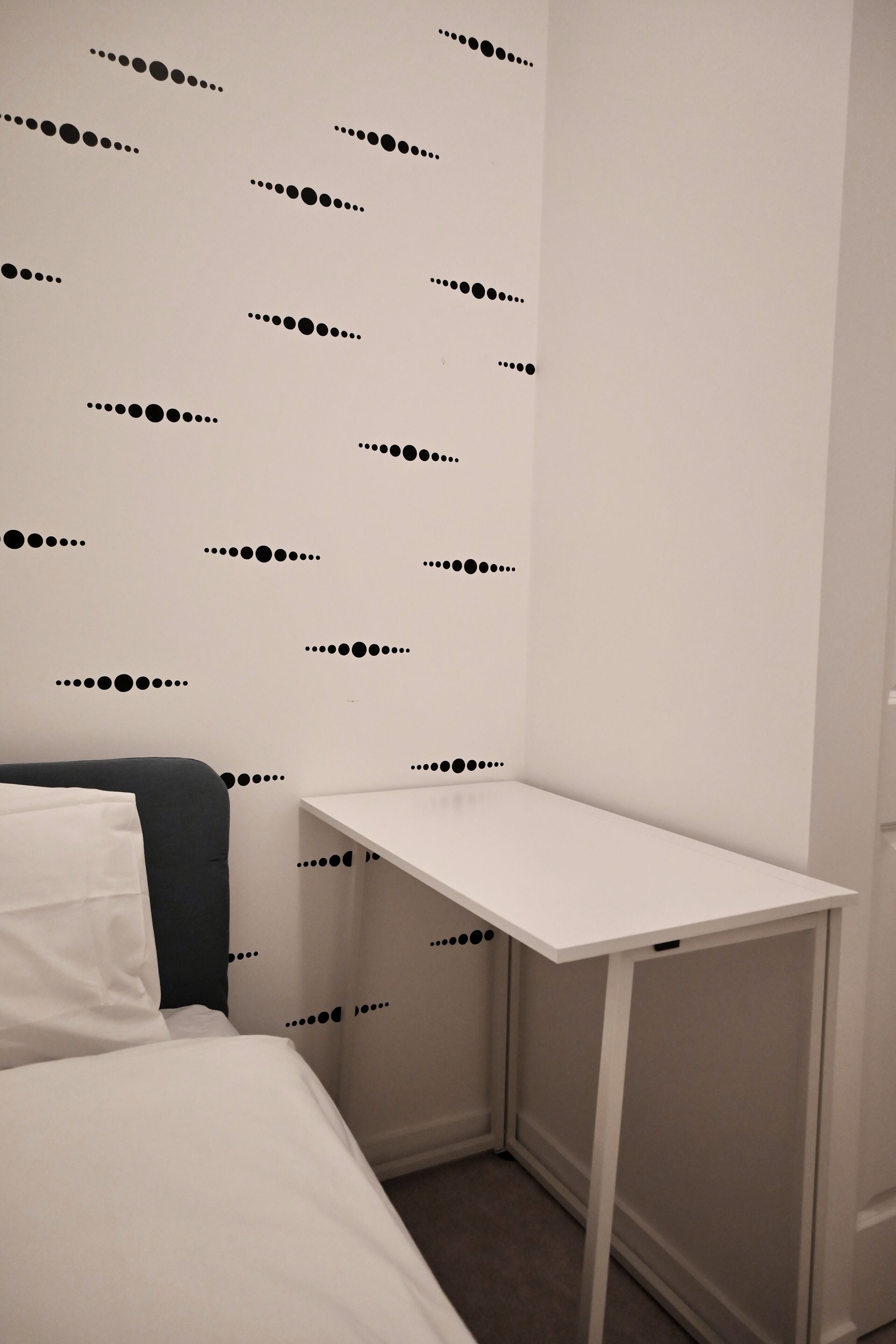 A small, neatly arranged bedroom with a single bed, a white desk, and a wall with a black dot pattern.