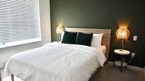 Ein ordentlich bezogenes Bett mit weißen Leinen, zwei Nachttischlampen und ein Fenster mit Jalousien.