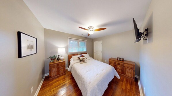 Une chambre à coucher avec un lit, des tables de chevet en bois, un ventilateur de plafond, une fenêtre avec des stores et une porte.