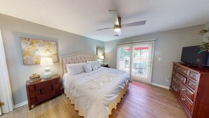 Une chambre à coucher comprenant un lit, une commode, une table de chevet, un ventilateur de plafond et une fenêtre avec des stores.